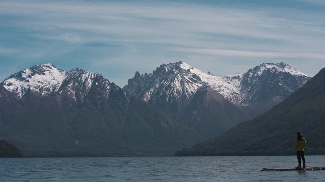 Man wearing a yellow jacket standing on the shore of Mascardi Lake, observing a dramatic mountain range with snow-capped peaks in Patagonia, Argentina.