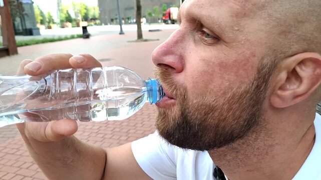 Side view of man holding plastic water bottle outdoors on city street, hydration and urban lifestyle in summer. Guy drinks water on a hot day
