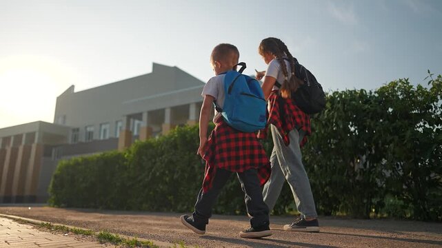 Student siblings walk along street in sunlight. Girl and boy walking hand in hand near school building. Morning road scene with backpack child walk. Student walk to school holding sibling hand