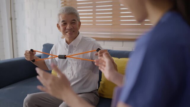 Health insurance with caregiver at home Senior man exercise band therapy rehabilitation caregiver home smile health living room support guided recovery sofa shoulder strength training indoors