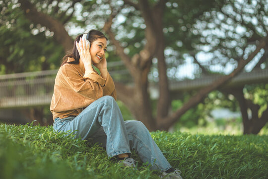 Happy Asian woman wearing headphones and smiling while sitting on green grass at park, young female listening to music, audio podcast, or streaming online in garden for modern digital lifestyle