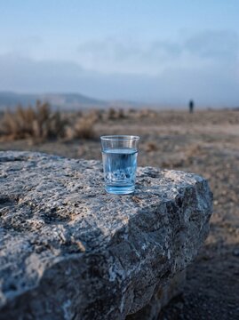 A clear glass of fresh water sitting on a rough stone in a vast desert landscape at dawn, symbolizing survival and hydration in an arid environment