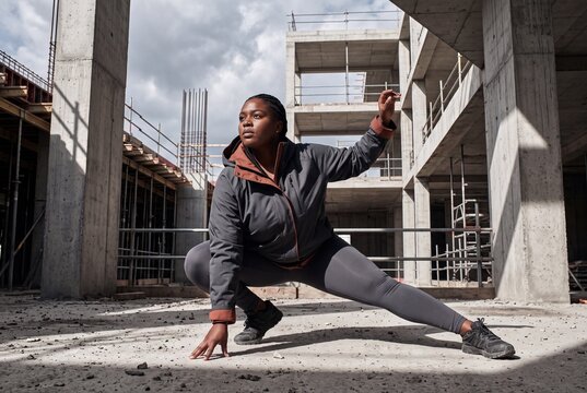 A strong Black woman performs a side lunge stretch inside an unfinished concrete building at a construction site, wearing athletic gear and looking focused