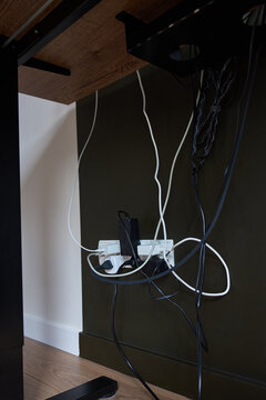 Tangled cables, power adapters and plugs connected to wall outlet under wooden desk