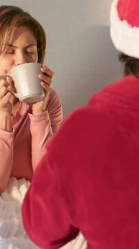 Vertical video: Sipping woman in pink top home holding mug, prompted by partner in red Santa hat
