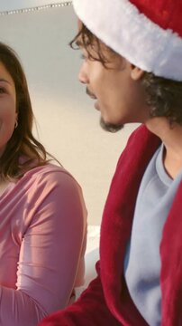Vertical video: Speaking woman starting holiday chat, man replying gesturing on bed with Santa hat