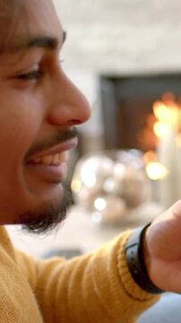 Vertical video: Smiling man in mustard knit checking phone and reacting near fireplace, wristband