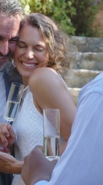 Vertical video: Leaning couple in suit and white dress holding flutes in yard, guest offering toast
