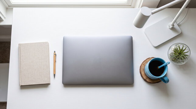 Minimalist workspace featuring a closed laptop a notebook and a blue coffee mug on a clean white desk surface lit by natural light from a window nearby