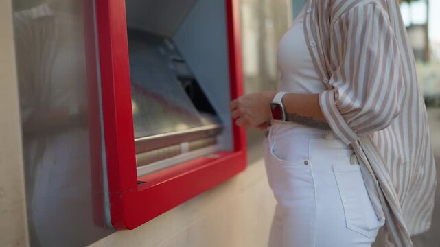 Unrecognizable woman standing outdoors while withdrawing money from a red atm, showcasing a common financial transaction and the convenience of modern banking technology on a city street
