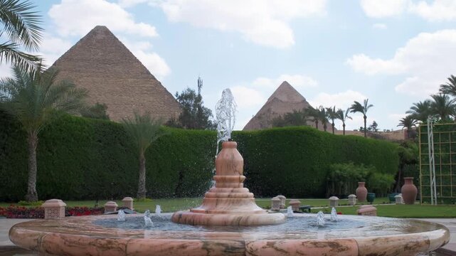 Slow motion 4K travel video featuring a fountain in the foreground with the Pyramids of Giza in the background, surrounded by palm trees and lush gardens in Egypt.