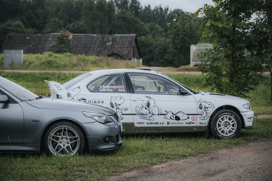 Madona, Latvia - July 11, 2026: Custom decorated racing car featuring Snoopy graphics parked beside a gray sports car in a rural outdoor setting with greenery