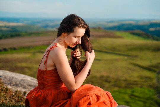 Woman sitting on a stone in a green field while playing with her long hair during a sunny afternoon
