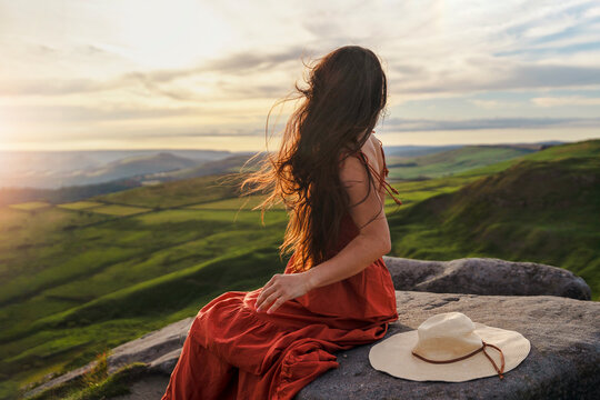 Woman sits on a rock and looks at the sunset over the hills with green fields in the background