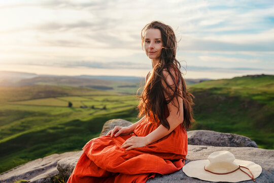 Woman sits on rocks wearing orange dress and straw hat, overlooking green hills during golden hour