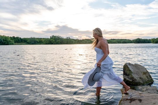 Woman walks into the water at lakeside during sunset while holding a hat and enjoying nature's beauty