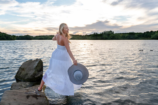 Woman walks by the water holding a large hat while looking back during sunset at a lake