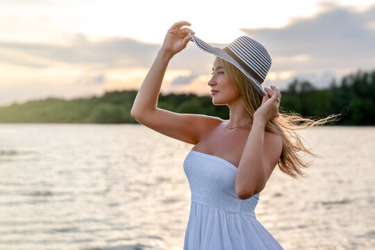 Woman wearing a hat stands by the water at sunset with trees in the background and clouds in the sky