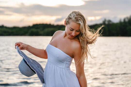 Woman stands by the water at sunset holding her hat while wearing a white dress and enjoying the warm evening air