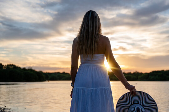 Woman stands by the water at sunset holding a hat while looking towards the horizon