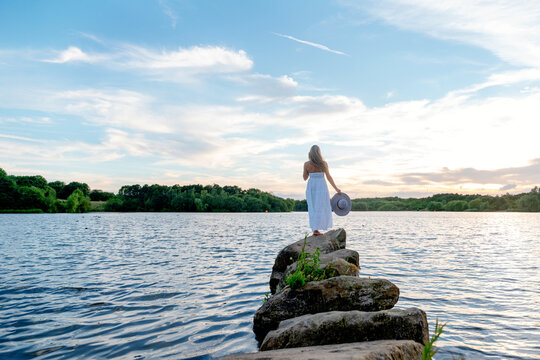 Woman standing on rocks at a lake during sunset with a hat in hand and trees in the background