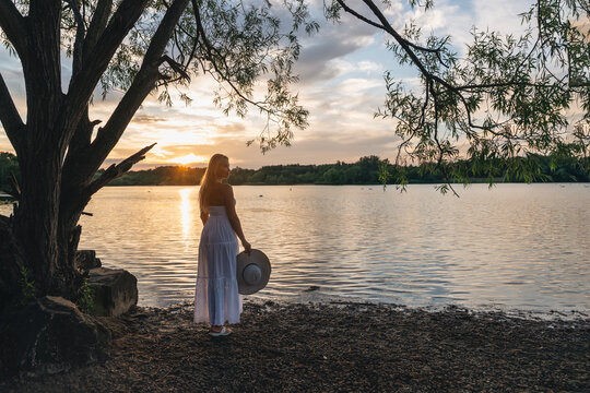Woman stands by the water during sunset wearing a white dress and a wide-brimmed hat near a lake surrounded by trees