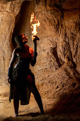 Naklejka premium Hooded warrior woman in fantasy costume holding a burning torch, exploring a dark rocky cave, dramatic fire light scene. Halloween theme