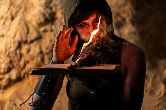 Mysterious hooded sorceress with red eyes casting a spell over a burning ancient book in a dark cave, dark fantasy magic and halloween scene