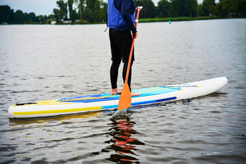 Person paddleboarding on a calm lake wearing a life jacket. Stand up paddleboarder performing a stroke with an orange paddle. Person standing on an inflatable SUP board in nature. © Rabizo Anatolii
