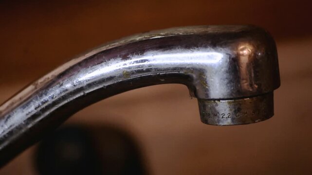 Close-up slow-motion footage of a leaky kitchen faucet with water dripping from the spout. Ideal for concepts of plumbing repairs, household maintenance, waste, and environmental awareness.