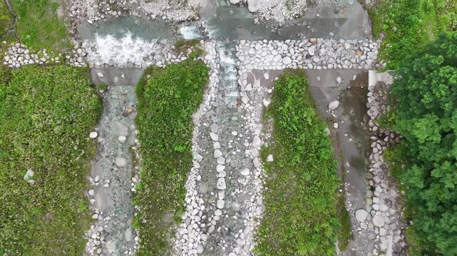 Aerial view of a winding river through rocky terrain. Water flows gently over stones and gravel. Green grass lines the riverbanks. Small path runs parallel to the water