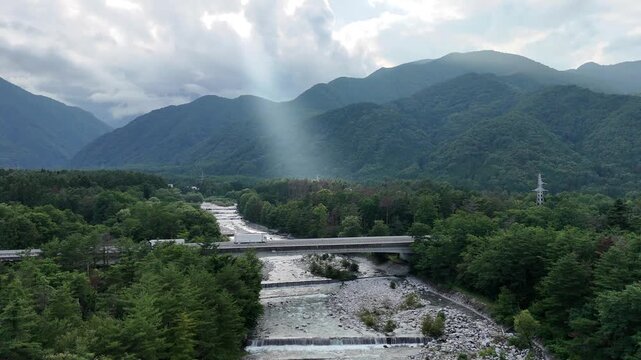 Mountain river winds through forested valley. Clouds part, letting rays light the scene. Bridge spans rocky stream, surrounded by green. Vast hills rise behind, lush and silent