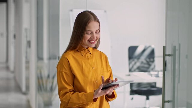 Smiling Young Caucasian Woman Engaged While Using Tablet In Modern Office. Happy Female Professional Holding Device, Working In Bright And Stylish Workspace, Excited About Task At Hand.