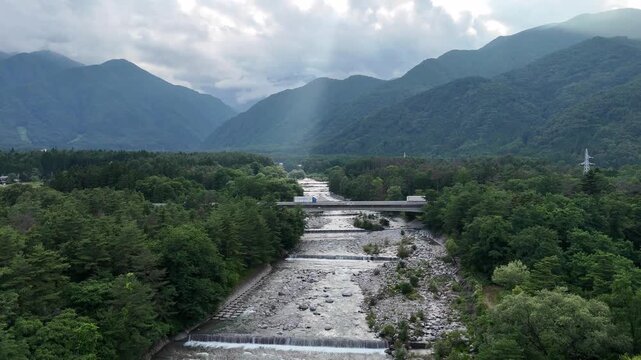 Riverside valley flows through dense forested mountains. Clouds cast soft light over misty peaks and tree-covered slopes. Stone bridge spans the rushing river, connecting green lands