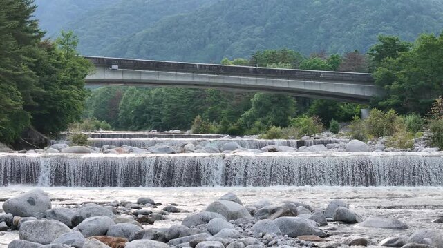 River flows over stones beneath a bridge. Green hills frame the scene with trees. Bridge spans the rushing water with calm grace. Nature and human-made structure coexist