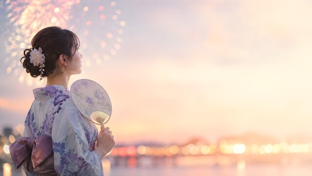 夕暮れに打ち上げ花火を眺めるうちわを持った浴衣姿の女性の横顔