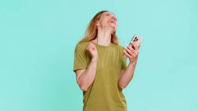 Middle-aged woman reads smartphone message and bursts into laughter, reacting with surprise and joy to unexpected compliment or lucky win. Adult girl isolated on blue background shows cheerful emotion