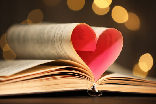 Book with heart made of pages on table against blurred lights, closeup