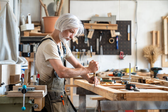 Qualified man carpenter working with vintage wooden door at workstation in restoration workshop, talented male restorer using chisel instrument for carving and cutting surface, detailed handmade work.