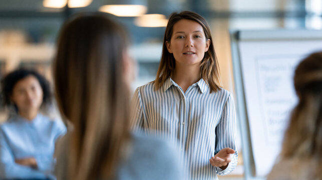Faceless female business coach presenting personal branding strategies on a flip chart to a small group in a bright studio, coaching and mentoring visible, defocused studio in