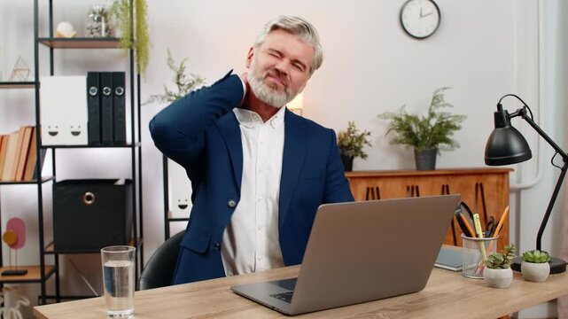 Middle-aged businessman at home office suddenly clutches neck feeling sharp deep pain grimacing needing urgent relief. Freelancer guy at table stretches muscles massaging area hoping discomfort eases.