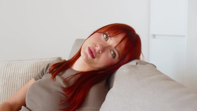 Young woman at home feels so exhausted and in bad mood after tough news stares at screen sighs softly. Redhead girl on sofa lowers gaze slow breath low energy no drive needs rest and calm before tasks