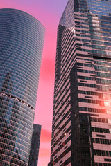 Fototapeta premium Towering financial office skyscrapers pierce colorful sunset-dawn sky vertical photo. Golden light washes over glass steel, casting reflections. skyscrapers shine business architecture.