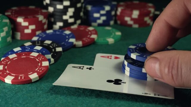 Hand placing blue poker chips on playing cards with aces, surrounded by colorful casino chips on a green felt table in a gaming environment