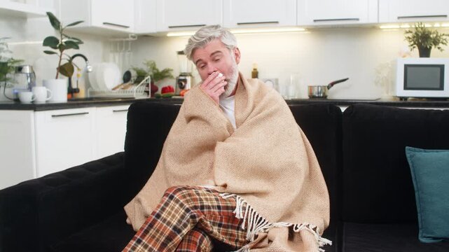 Middle-aged man at home sofa under plaid coughs wipes nose with tissue chills and sniffles during a hard flu, sneezes wipes snot into napkin feels cold suffering from allergy flu symptoms quarantine.
