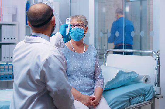 Doctor checks senior patient temperature in clinic with thermometer screening, protective equipment and health safety workflow during a private clinic appointment.