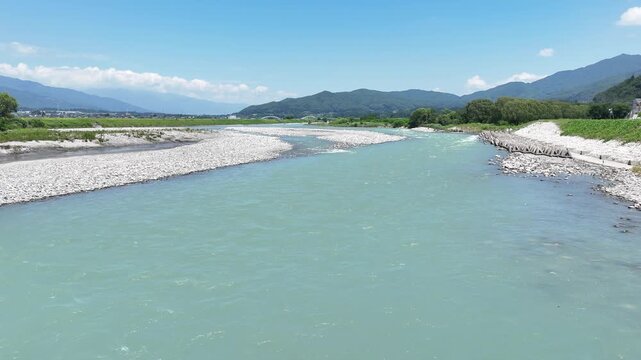 Clear river flows through rocky bed. Sky above is bright and blue. Mountains frame the distant horizon. Riverbed shows smooth stones and gravel. Water moves gently, reflecting sky