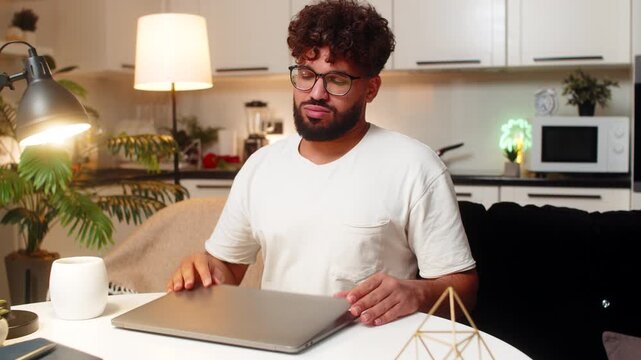 Arabian man at home closes laptop after tasks exhales relaxes breath slows remote work done. Egyptian guy sitting at table looks satisfied rubs eyes stretches shoulders enjoys short break tonight.