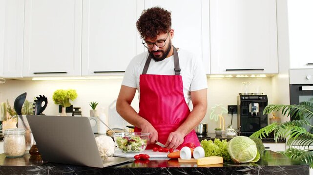 Arabian man prepares fresh salad in kitchen following recipe on laptop for healthy diet clean eating and meal prep. Young guy feels focused and confident chopping vegetables at home apartment indoors.