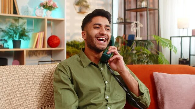 Indian man on home sofa talks on wired phone with joy during a happy call about good news or family support. Arabian guy on home sofa laughs during friendly conversation showing joy relief and trust.